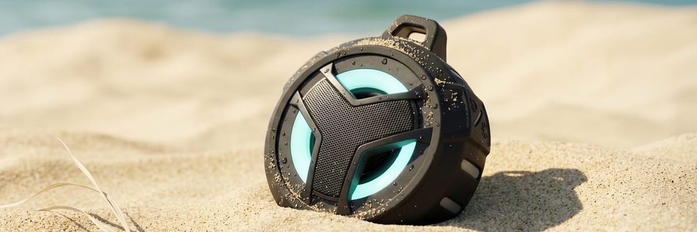EBODA Bluetooth shower speaker, a matte-black rugged cube with a cyan LED ring, resting in sand on a sunlit beach