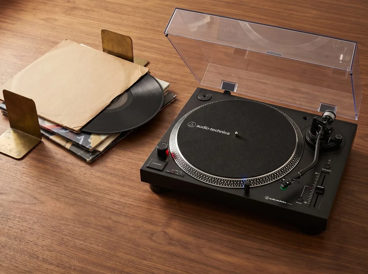 Audio-Technica AT-LP120XUSB turntable on walnut beside a leaning stack of vinyl records in kraft sleeves